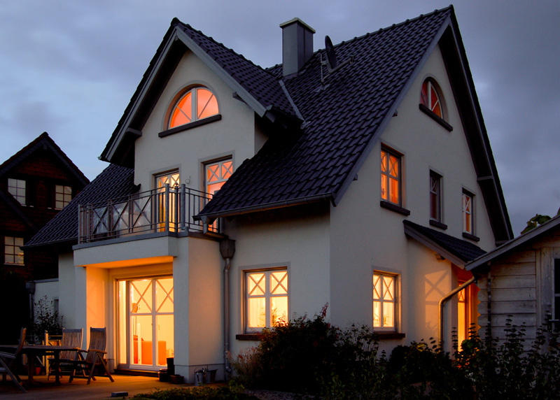 Weißes Einfamilienhaus mit Rundbogenfenster und Balkon, abends beleuchtete Fenster