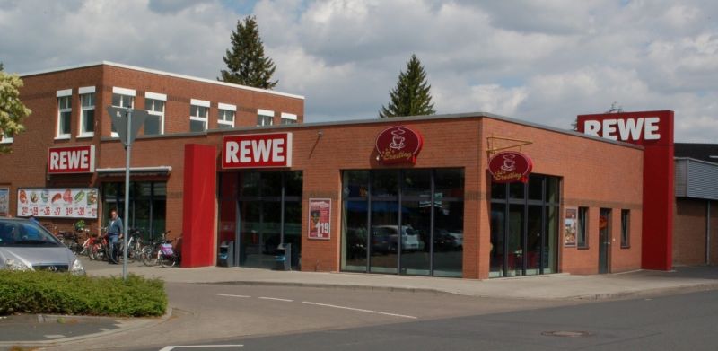 REWE-Markt mit Klinkerfassade, roten Eingangsportalen und Bäckerei an der Straßenecke